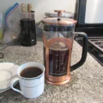 Copper French press next to a cup of brewed coffee on a kitchen counter